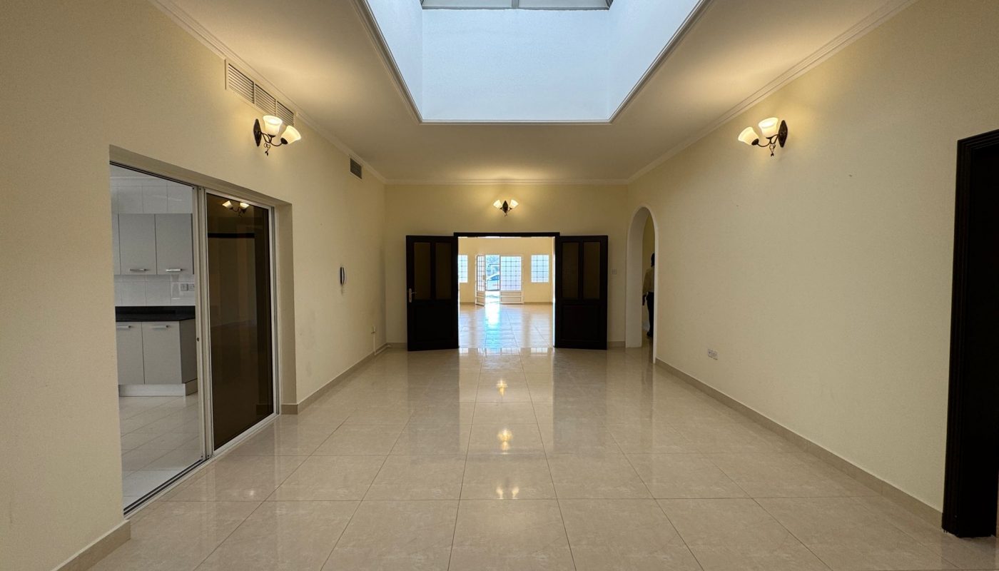 Unfurnished room with tiled floor, cream-colored walls, a skylight, wall sconces, and a view into a kitchen on the left and another room straight ahead through double doors.