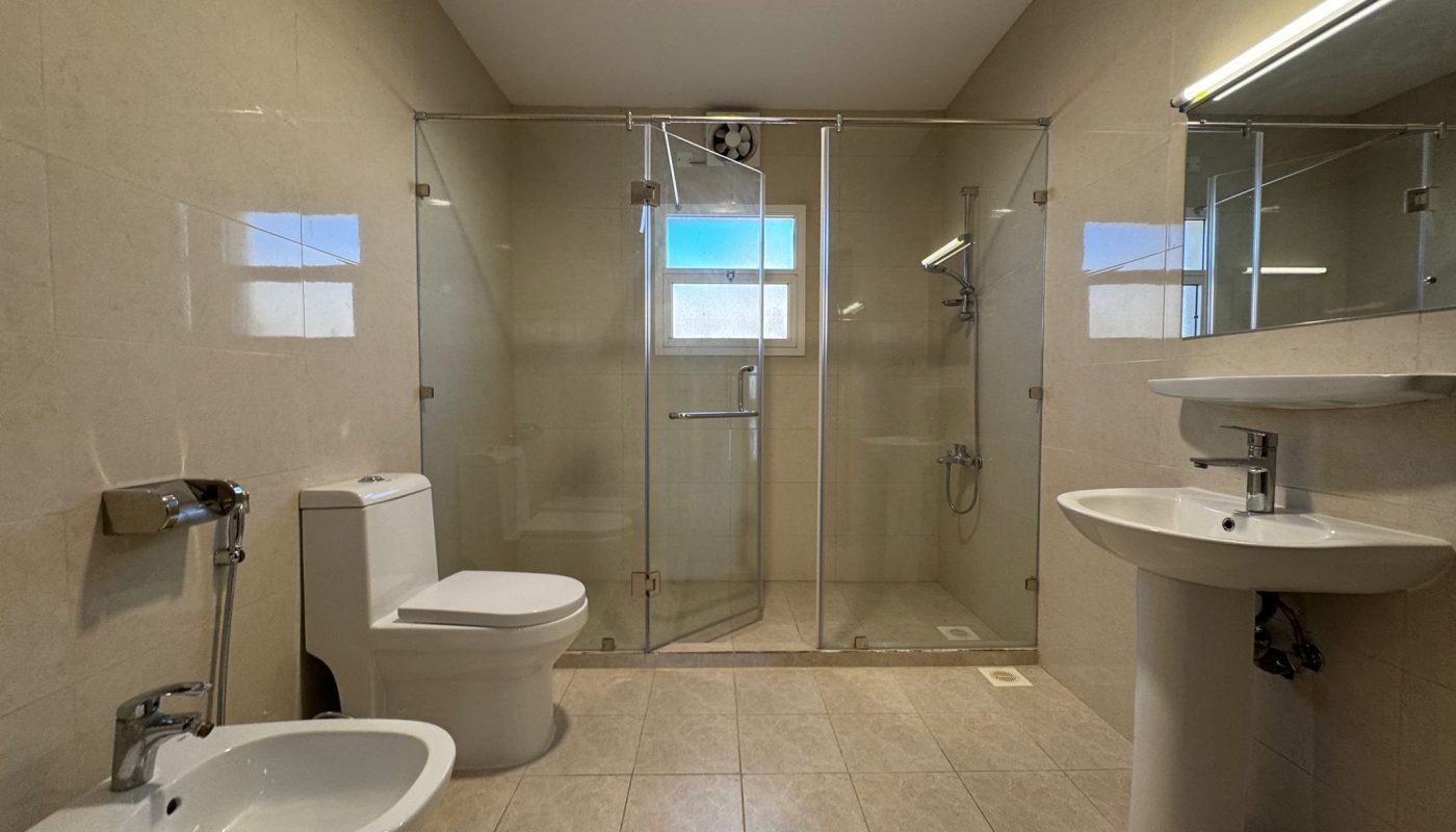Modern bathroom with beige tiles, a toilet, bidet, two sinks, and a glass-enclosed shower area with a window above.