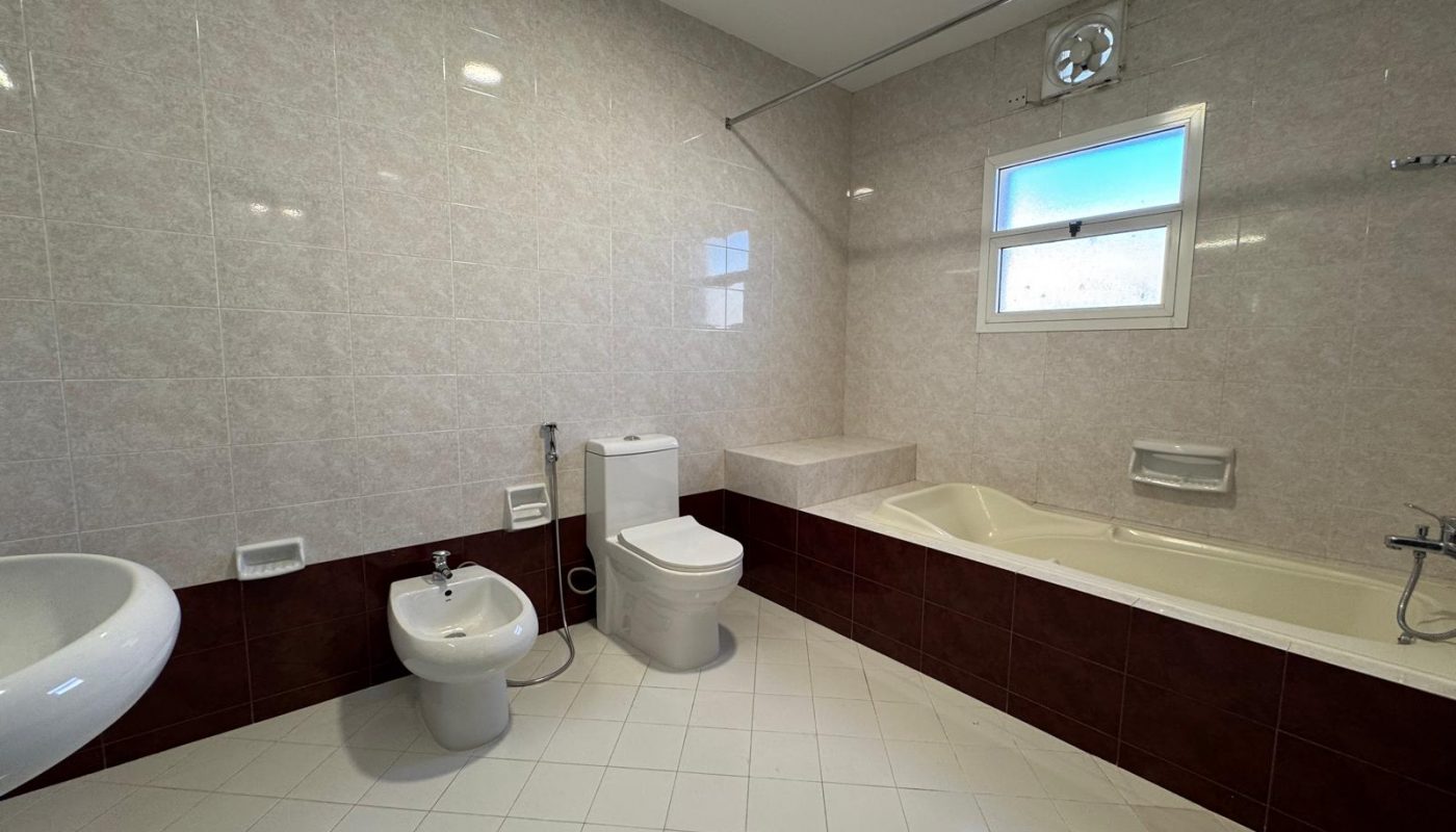 A bathroom with a sink, bidet, toilet, large bathtub, tiled walls and floor, a window, and a wall-mounted exhaust fan.