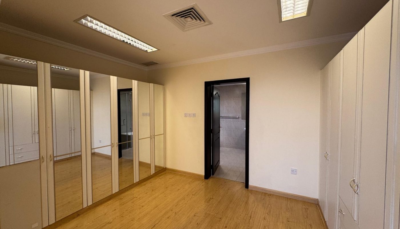 Empty room with wooden floor, built-in wardrobes with mirrors, white cabinets, and a doorway leading to a tiled bathroom. Ceiling has recessed lighting and air vent.