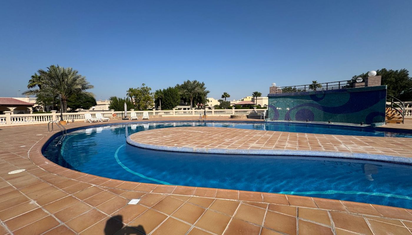 Outdoor swimming pool with clear blue water, surrounded by a tiled deck, lounge chairs, and palm trees under a clear sky. A person's shadow is visible in the foreground.