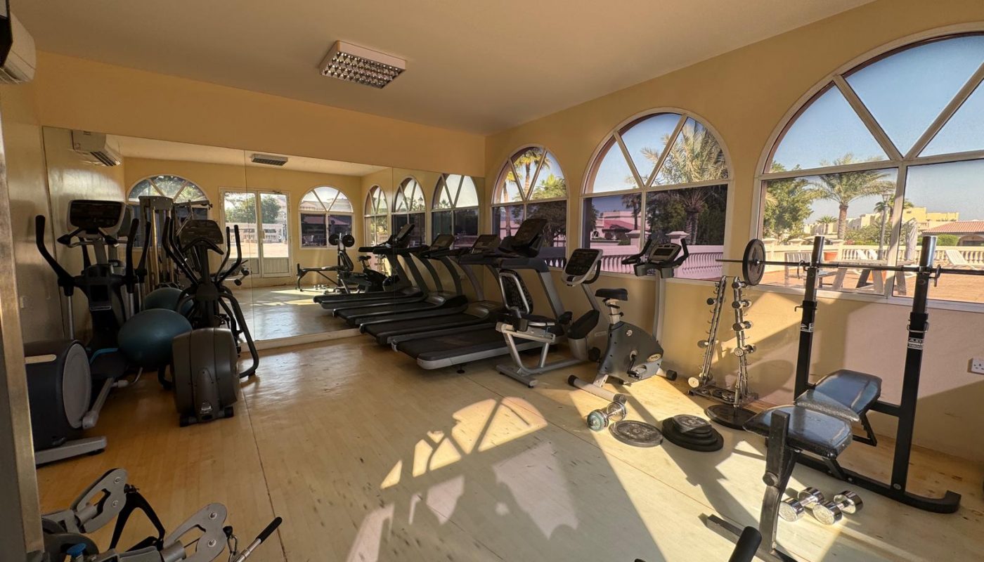 A gym room with treadmills, ellipticals, exercise bikes, weight machines, free weights, mirrors, and arched windows letting in sunlight.