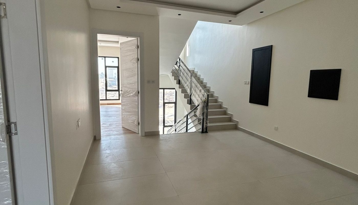 Spacious, modern hallway with light-colored tiled floor, white walls, and a staircase leading upstairs; large windows provide natural light.