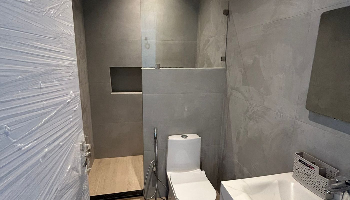 Modern bathroom with a toilet, bidet spray, wall-mounted sink, mirror, and a glass-enclosed shower area. Neutral gray walls and light wood-style flooring.