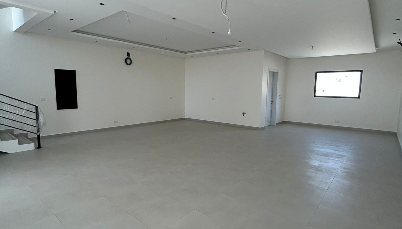 Spacious, empty room with white walls, light gray tiled floor, a staircase on the left, a window on the right, and unfinished ceiling fixtures.
