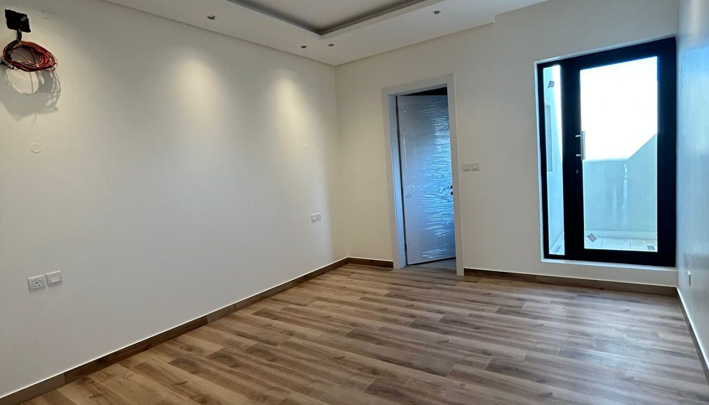 Empty room with wood flooring, white walls, recessed ceiling lights, a doorway with a closed door, and a glass door leading to an outside balcony.