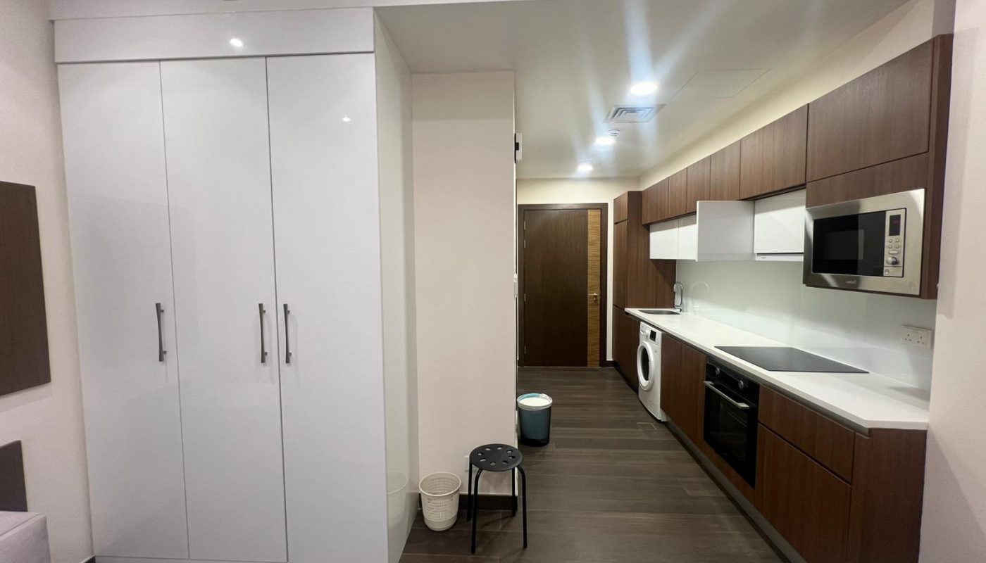 Modern kitchen with white and wood cabinets, built-in appliances, a microwave, washing machine, stovetop, sink, and storage cupboards, with dark flooring and neutral walls.