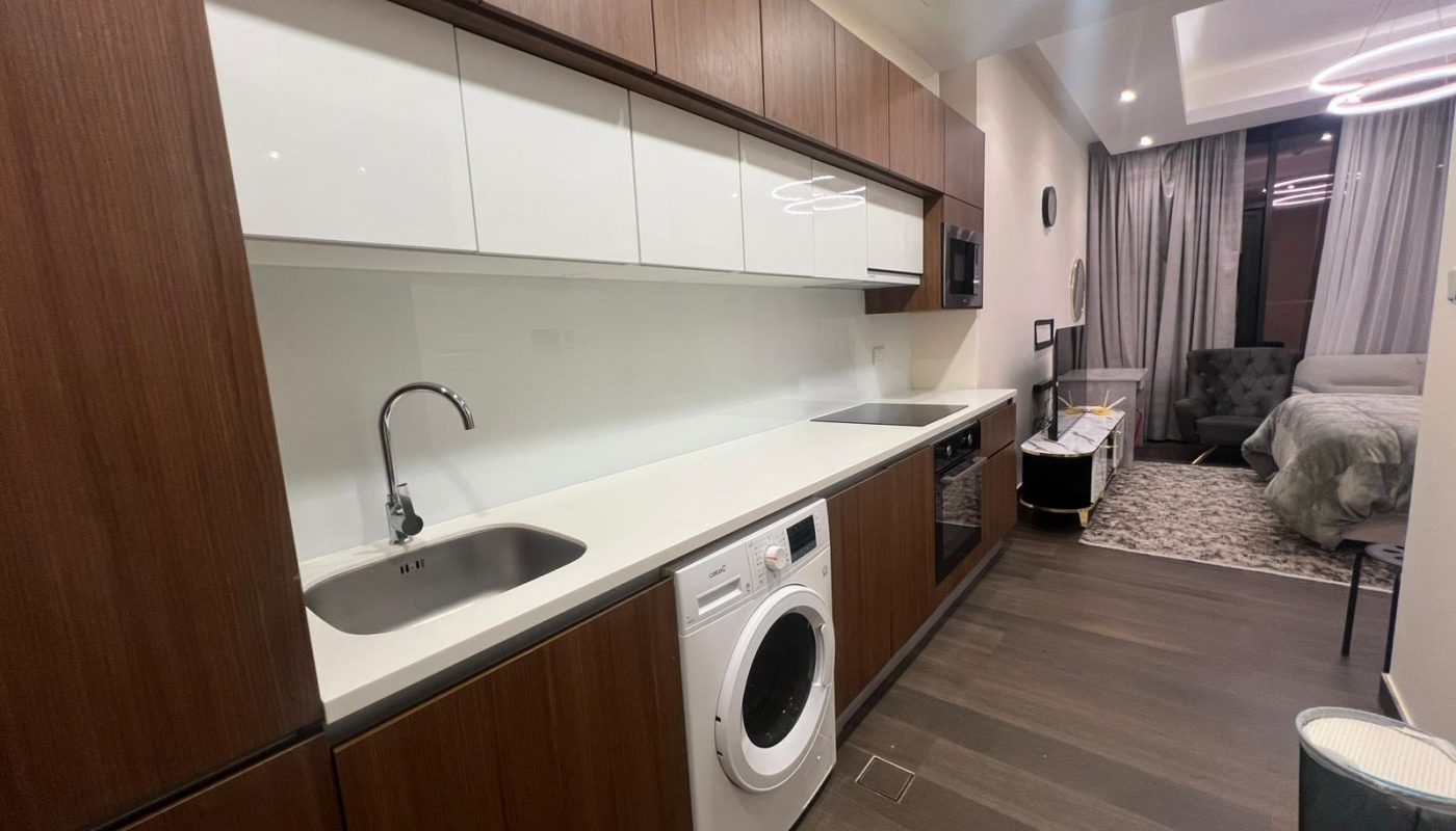 Modern studio apartment kitchenette with wood cabinets, white countertops, built-in appliances, and a washing machine; open layout extends to a living and sleeping area.