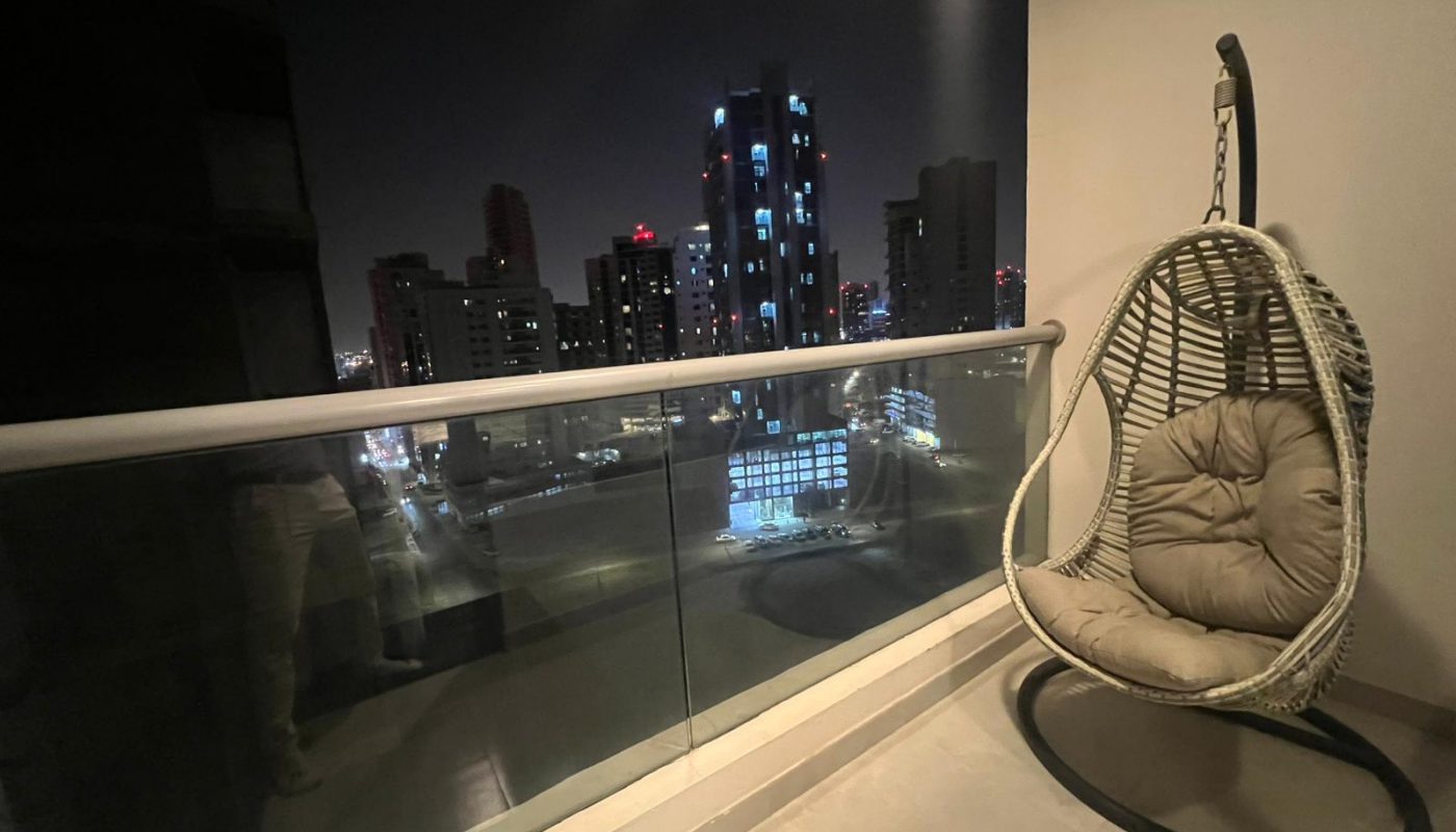 A balcony at night with a hanging wicker chair and cushion, overlooking city buildings illuminated by lights.