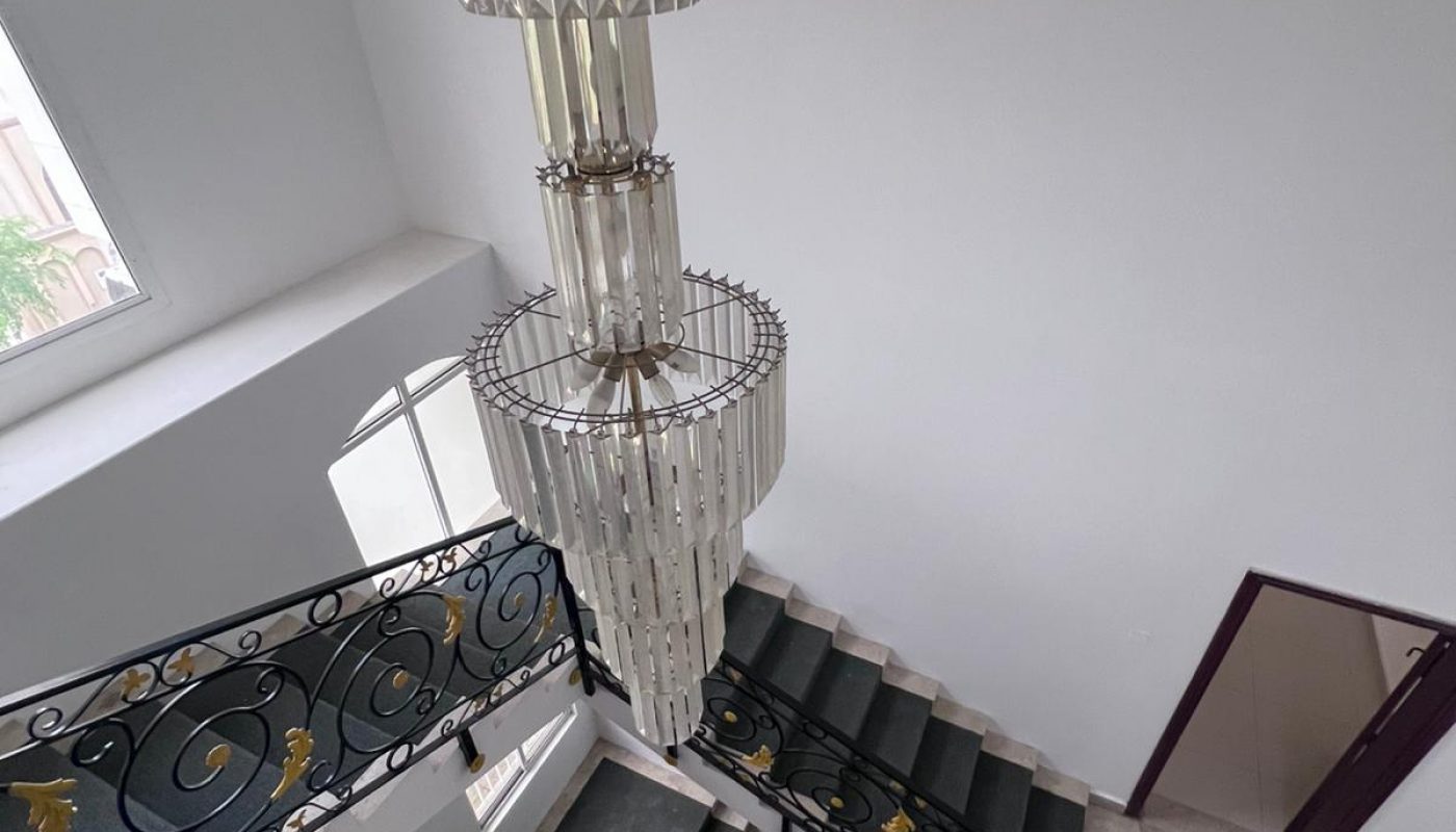 A large glass chandelier hangs above a black and white staircase with ornate railings in a bright, white-walled interior.