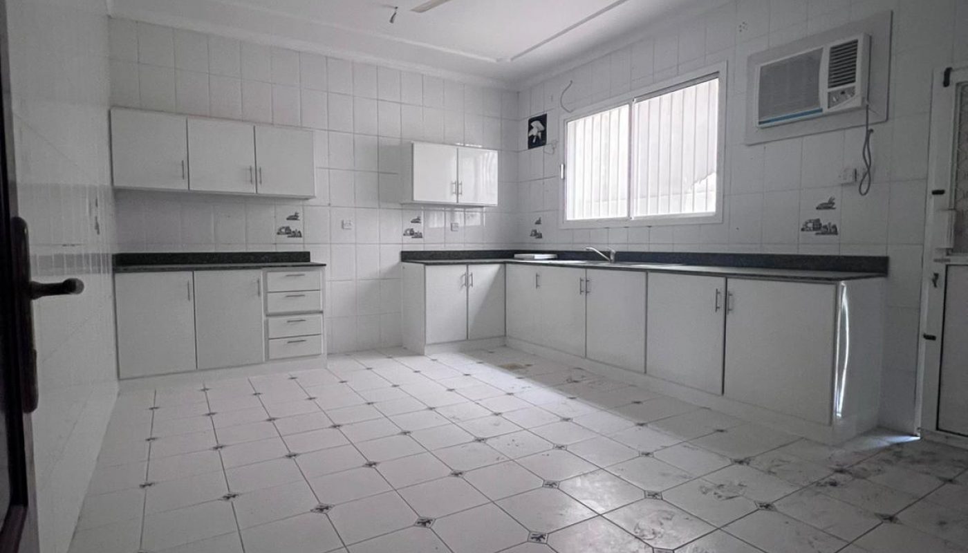 Empty kitchen with white tiled floors and walls, white cabinets, a ceiling fan, a window, and a wall-mounted air conditioner. Some dirt is visible on the floor.