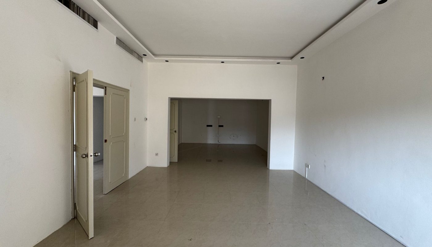 Empty, unfurnished room with white walls, beige tiled floor, an open door on the left, and a large rectangular opening at the back leading to another space.