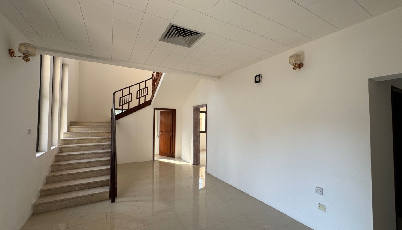 Empty room with tiled floor, white walls, wall-mounted light fixtures, a staircase with a dark handrail, and a wooden door under natural light.