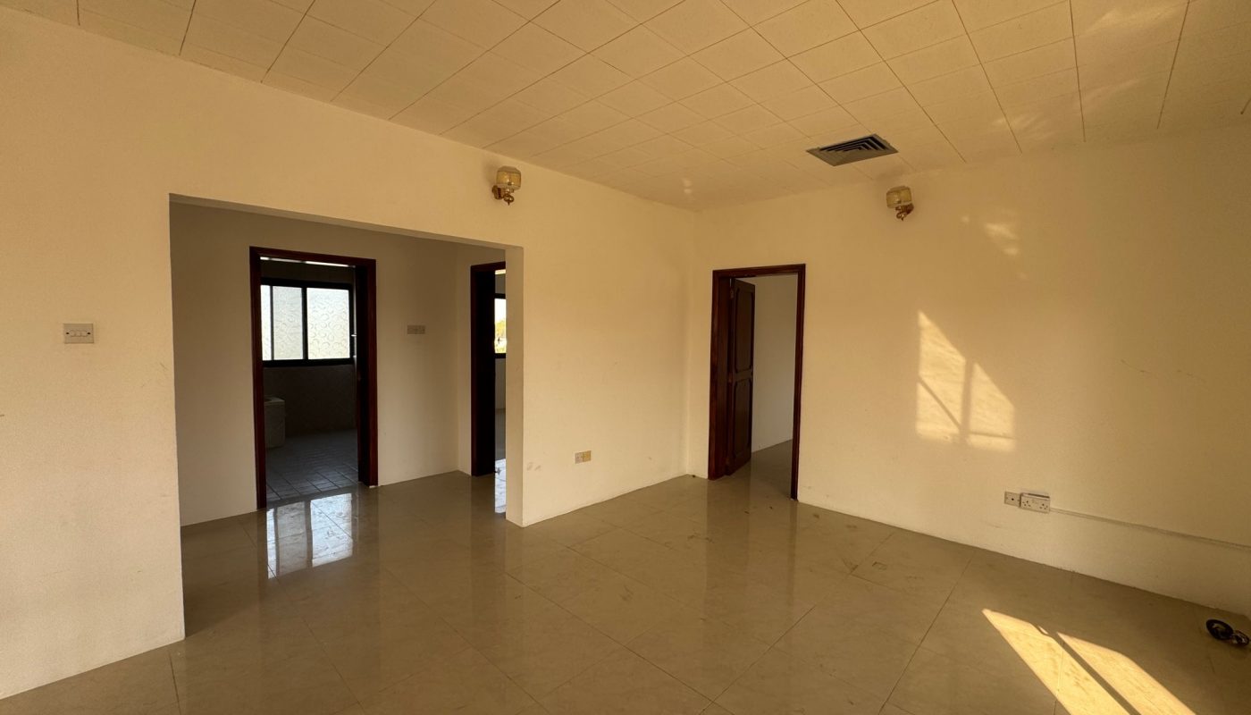 Empty room with tiled floor, cream-colored walls, ceiling lights, two doorways, one closed wooden door, and sunlight streaming through a window.
