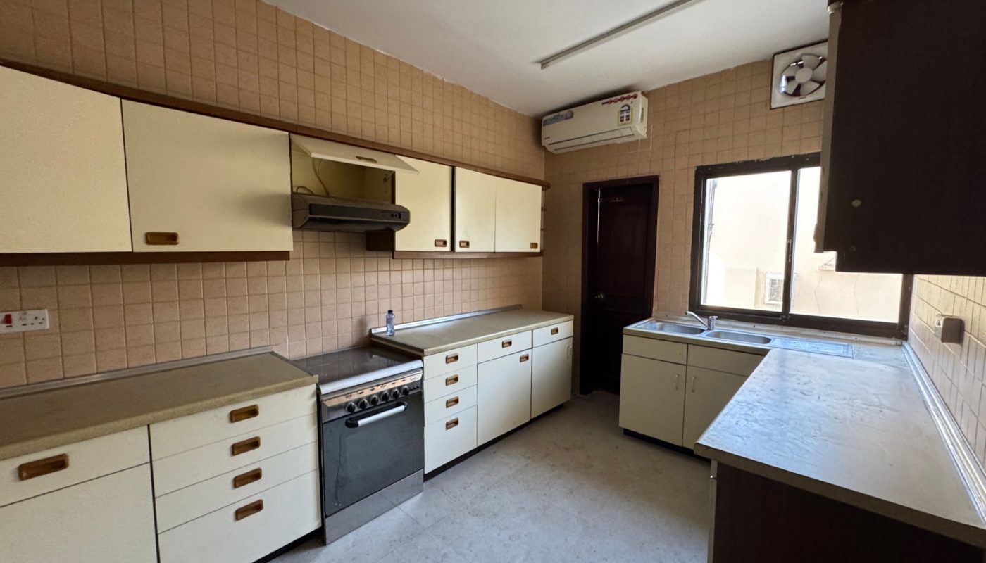 A simple kitchen with cream-colored cabinets, a stove with a range hood, countertops, a sink below a window, and an air conditioner on the wall.