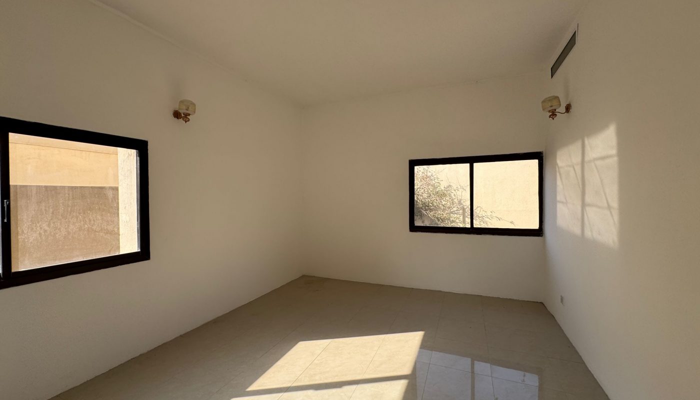 Empty room with white walls, tiled floor, two windows letting in sunlight, and two wall-mounted light fixtures. Shadows from the window frame are visible on the floor and wall.