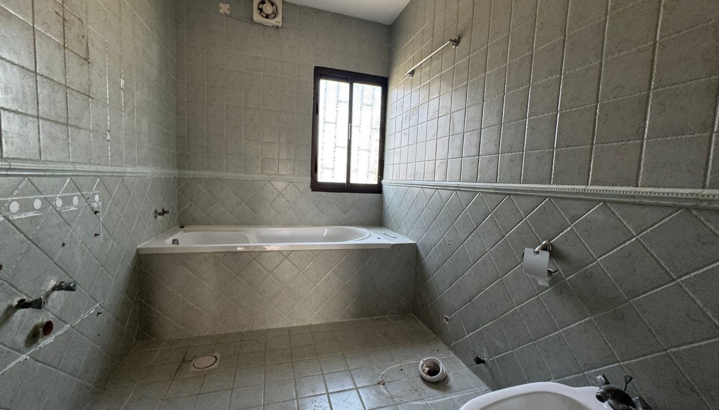 Empty bathroom with greenish tiles, a bathtub under a window, exposed plumbing, wall vent, and a sink in the foreground. Fixtures and fittings are missing.