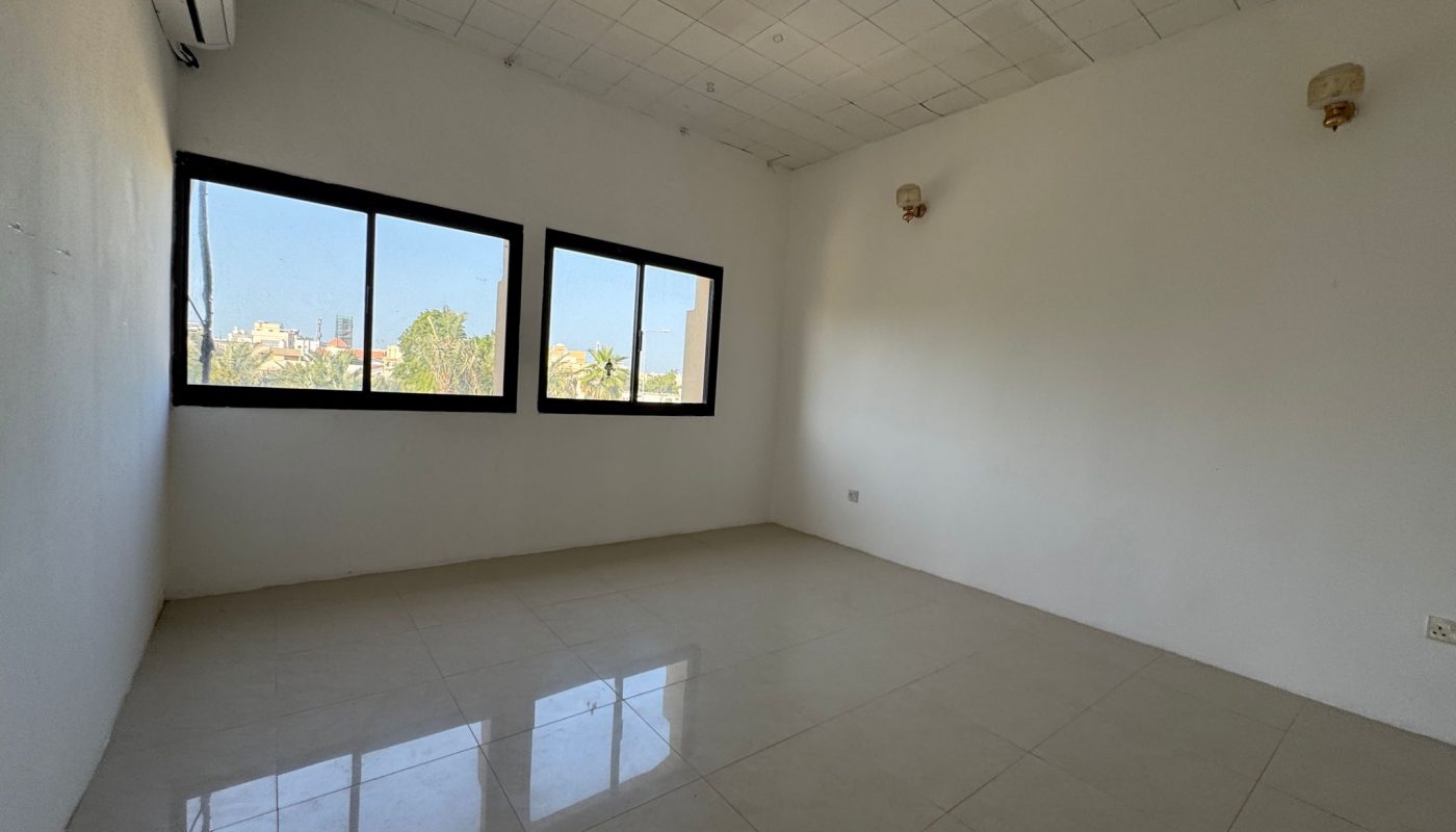 Empty room with white walls, tiled floor, three windows showing trees and buildings outside, two wall-mounted lights, and an air conditioning unit.