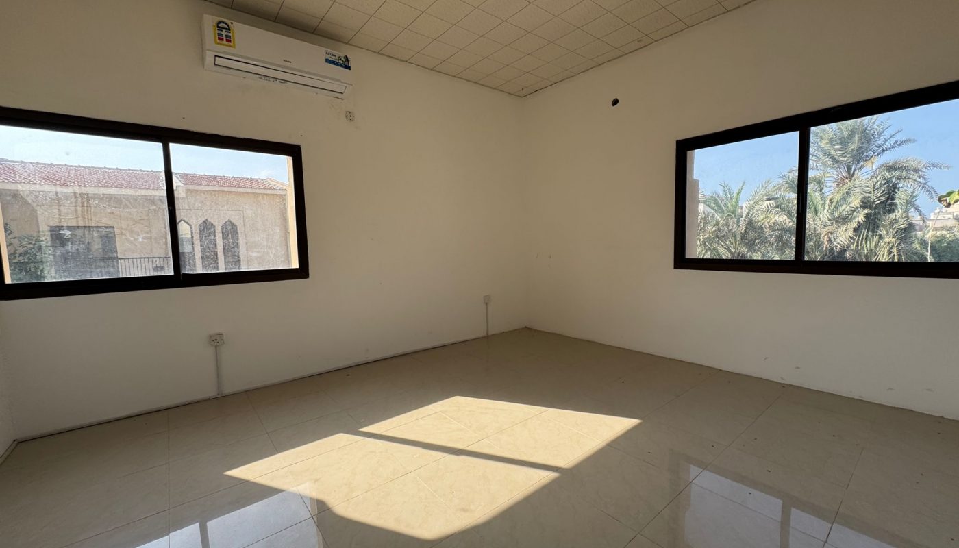 Empty room with tiled floor, two large windows, and an air conditioning unit mounted on the wall. Sunlight is casting a square shadow on the floor.