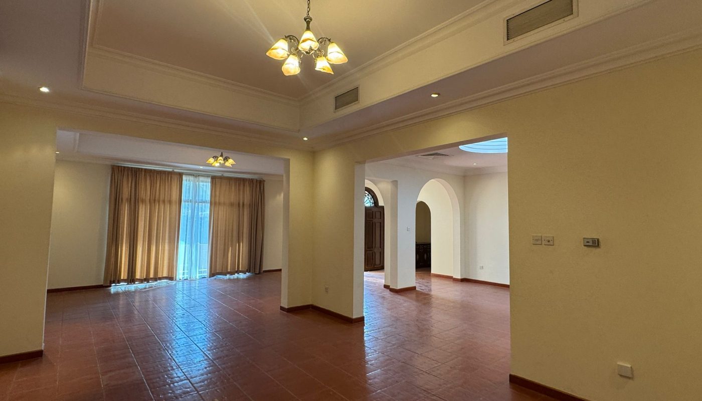 Spacious empty living room with tiled floor, cream-colored walls, ceiling lights, large windows with curtains, and an arched doorway leading to another room.