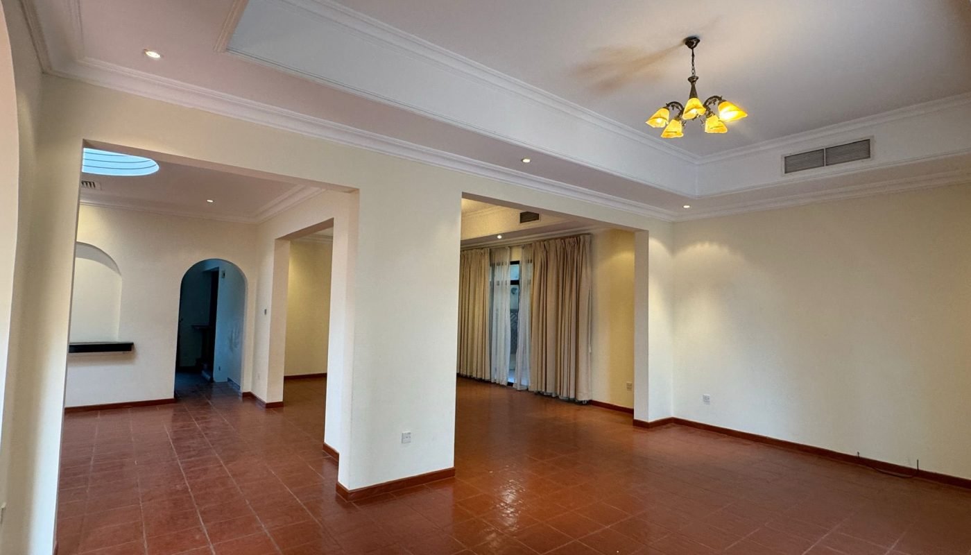 Spacious empty living room with terracotta floor tiles, cream-colored walls, ceiling lights, large windows with curtains, and arched doorways.