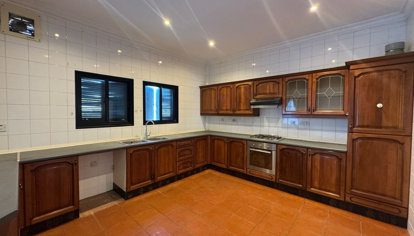 Spacious kitchen with brown wooden cabinets, white tiled walls, orange tiled floor, black-framed windows, and built-in stove with oven. Ceiling spotlights illuminate the room.