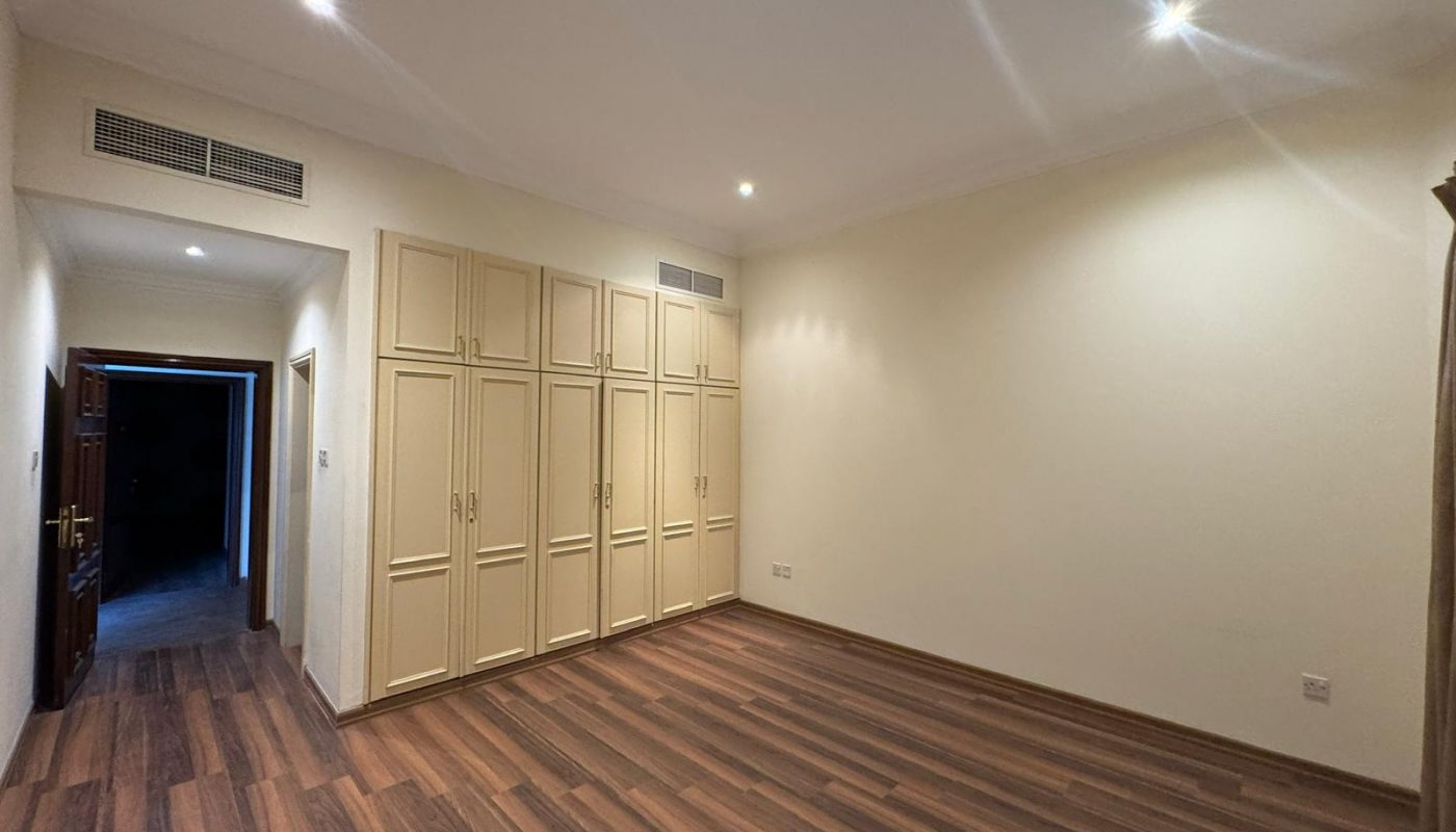 Empty room with wooden flooring, cream-colored walls, built-in cream wardrobes, recessed ceiling lights, and an open doorway leading to a darker hall.