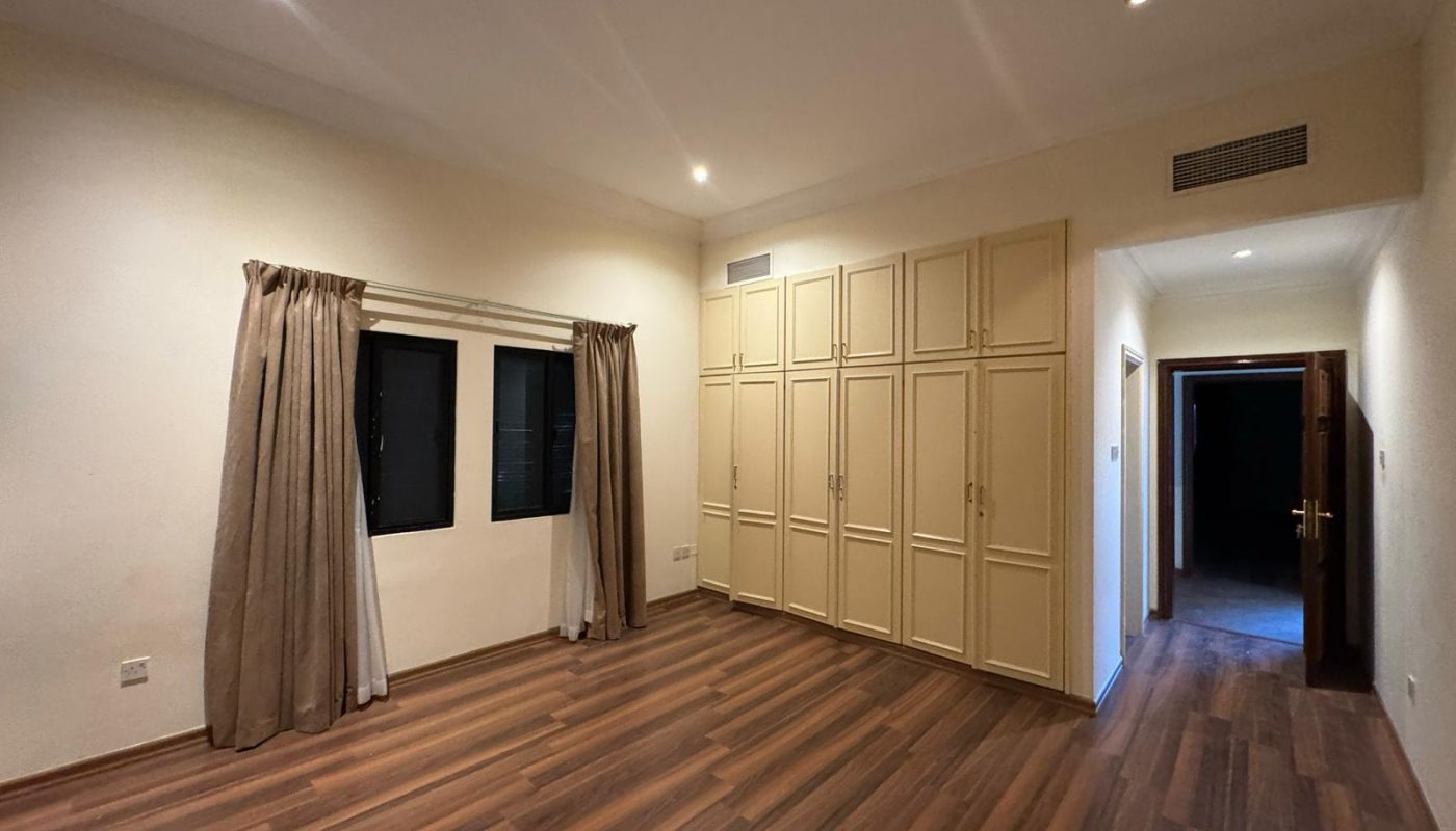 Empty room with wood flooring, beige built-in wardrobes, two windows with brown curtains, cream walls, and recessed ceiling lights. An open doorway leads to a darker room.