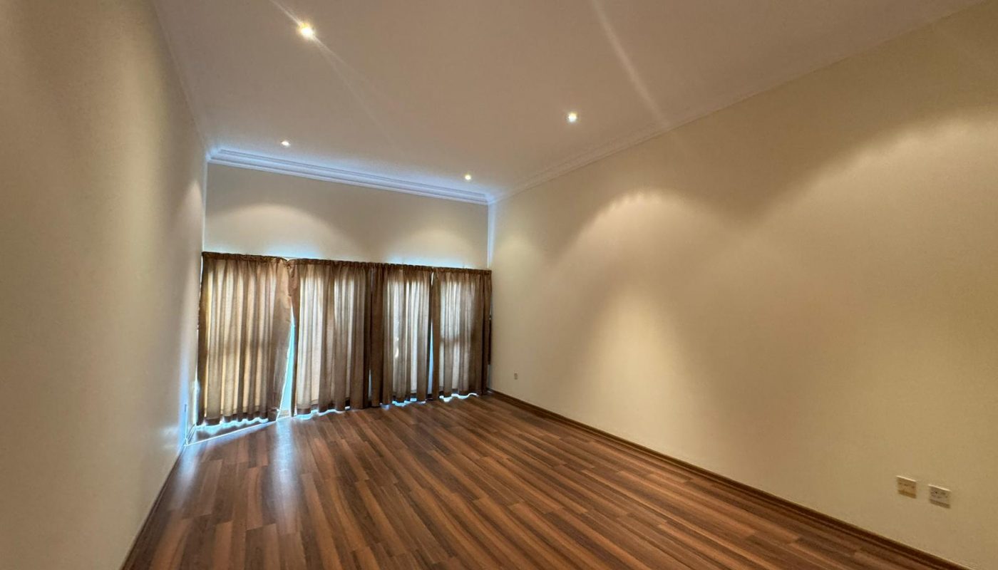 Empty room with wood flooring, cream-colored walls, ceiling spotlights, and large windows covered by brown curtains.