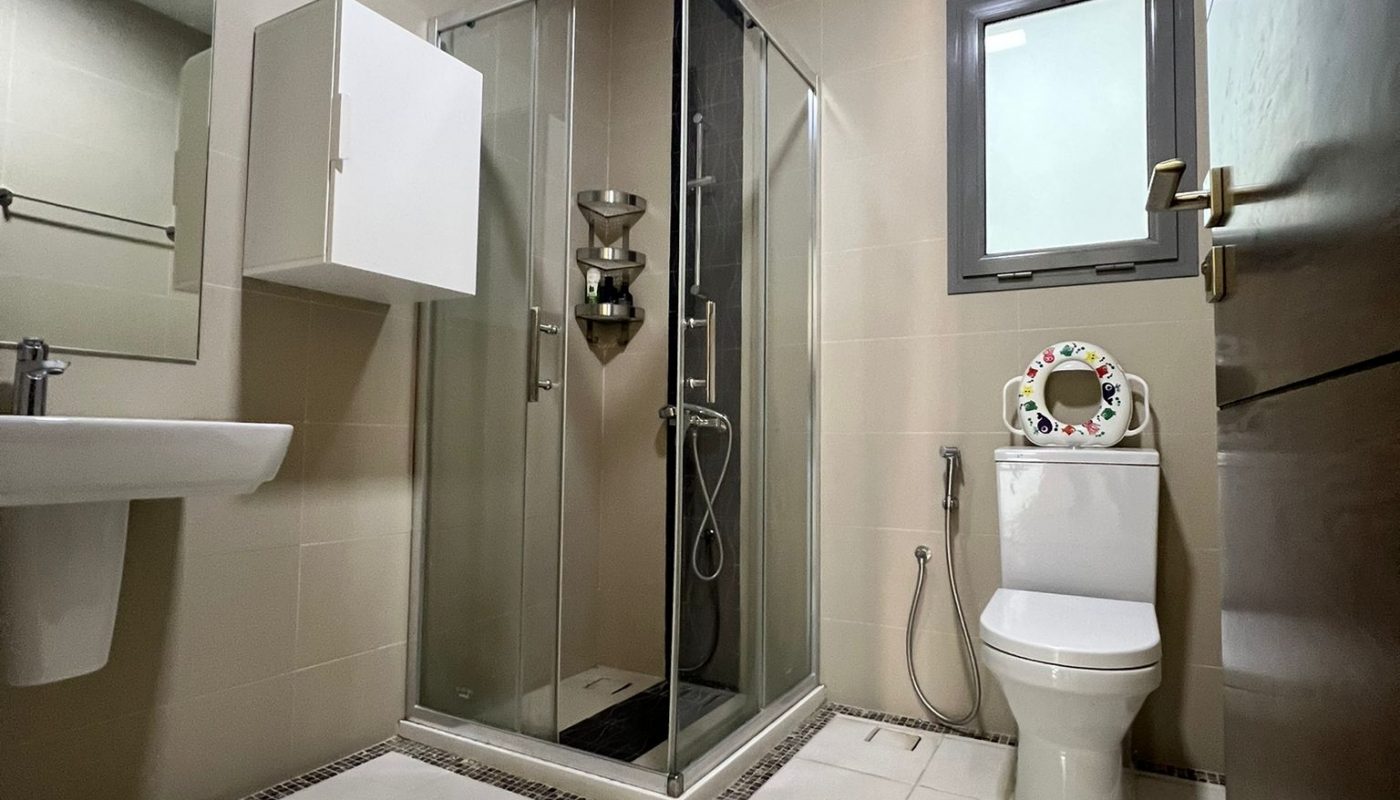 A modern bathroom with a glass shower enclosure, a wall-mounted sink, a toilet with a bidet spray, a mirror, and beige tiled walls and floors.