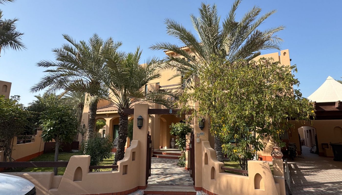 A two-story beige villa with palm trees and landscaped greenery in the front yard, seen under a clear blue sky. A black car is partially visible on the left side.