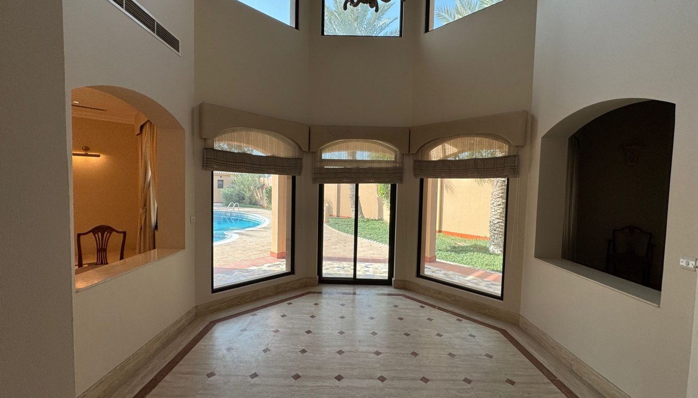 A spacious, empty room with tall windows, marble flooring, and a view of a backyard pool. A chandelier hangs from the ceiling and a dining area is visible on the left.