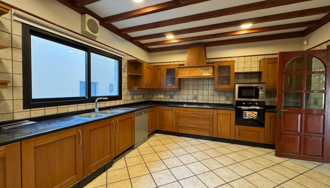 Spacious kitchen with wooden cabinets, black countertops, tiled floor and walls, large window, stainless steel appliances, and open wooden door.