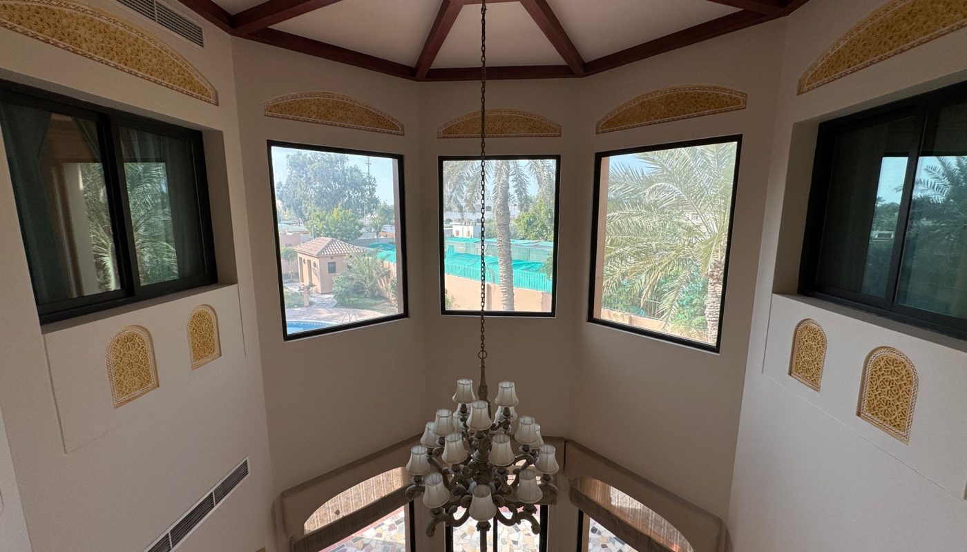 A view of a high-ceilinged room with large windows, a chandelier, geometric floor tiles, decorative wall accents, and visible greenery outside.