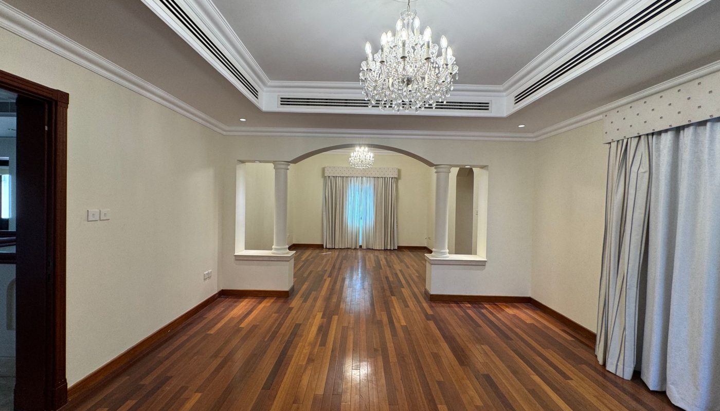 Spacious, empty living room with wooden flooring, two chandeliers, cream-colored walls, and large windows covered with curtains.