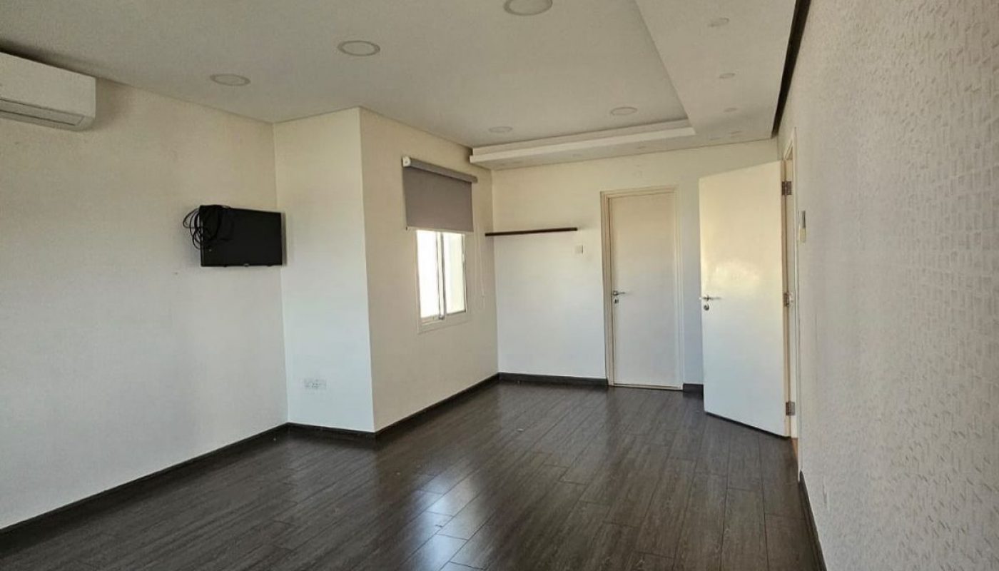 Empty room with dark wood flooring, white walls, a closed white door, a window with a gray blind, wall-mounted TV, and air conditioner unit.