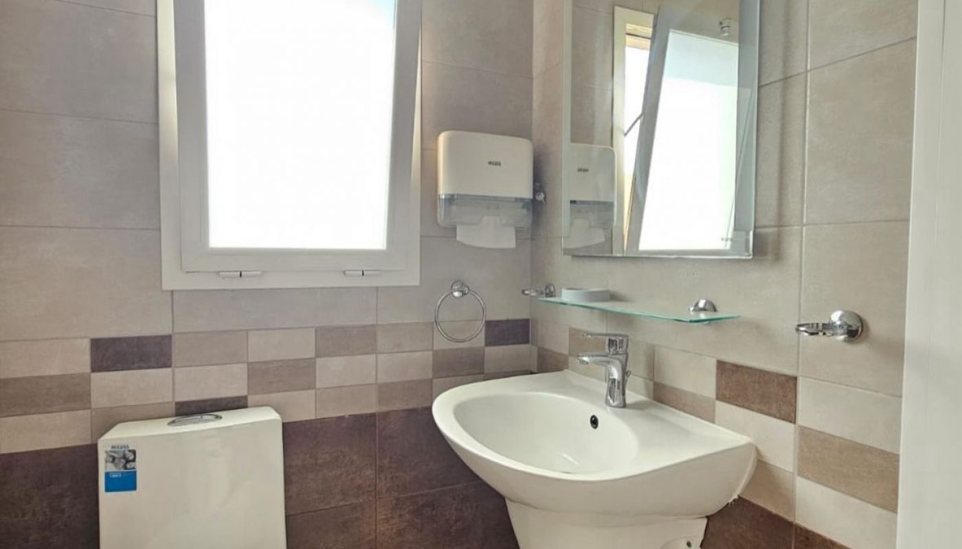Modern bathroom with a wall-mounted sink, toilet, mirror, paper towel dispenser, towel ring, window, and small black trash bin. Beige and brown tiled walls and floor.