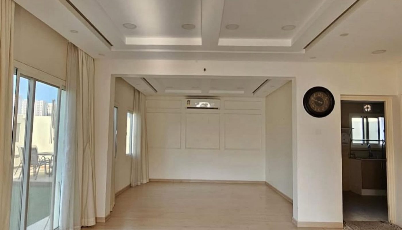Spacious, empty room with light wood floor, white walls and ceiling, large windows with curtains, and a small kitchen visible on the right.