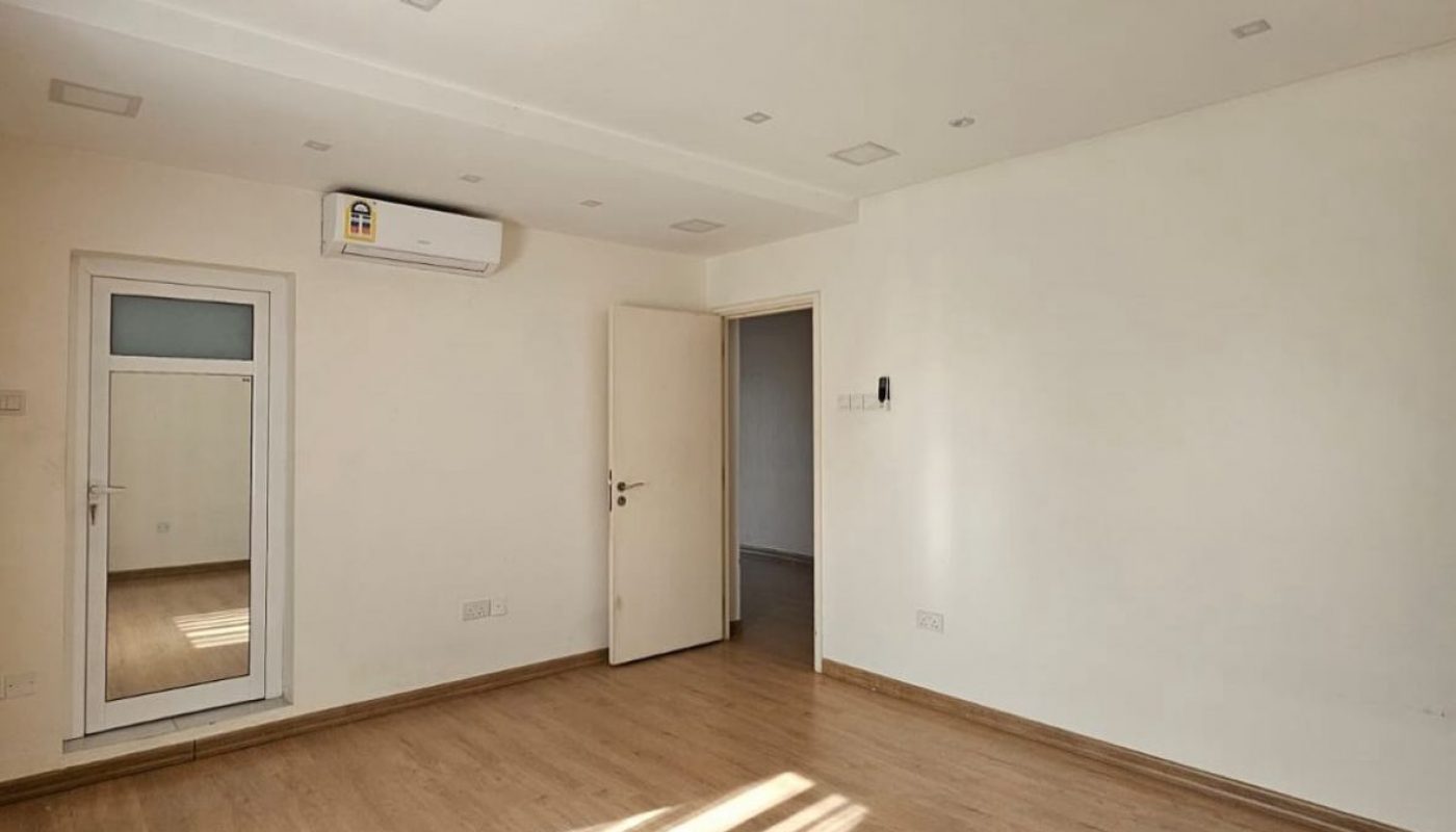Empty room with white walls, wooden flooring, an open door, a glass door, an air conditioner, and sunlight streaming through a window.