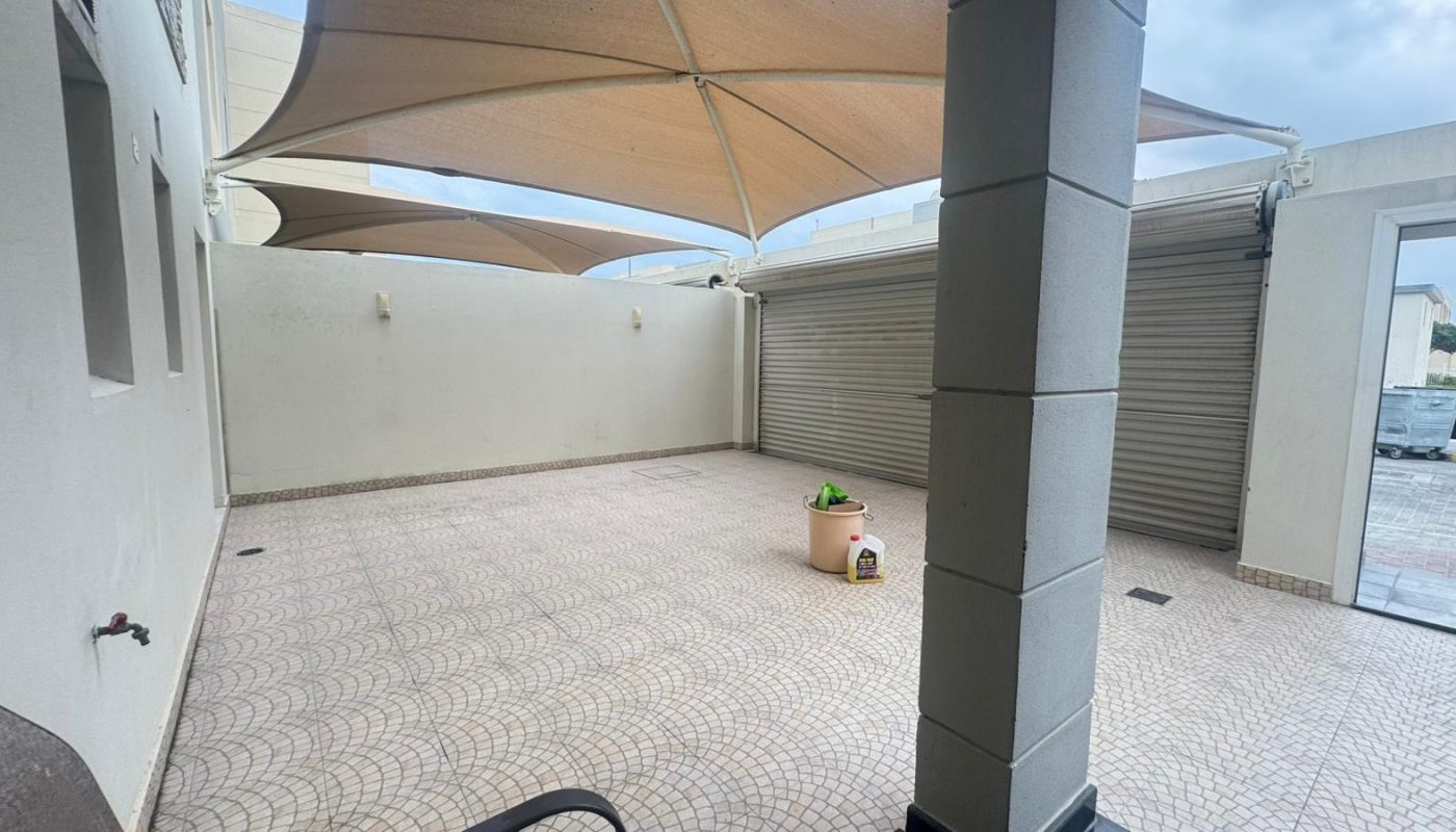 A tiled outdoor patio with a shade canopy, two beige walls, a large closed metal garage door, and a few cleaning supplies on the ground.