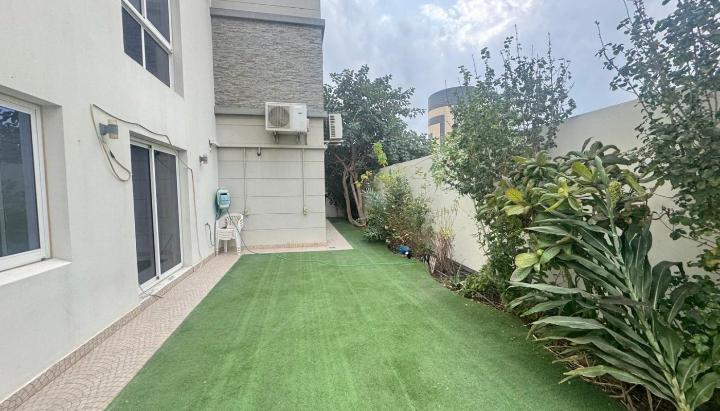 Side view of a modern house with artificial grass lawn, potted plants, outdoor chairs, and a wall-mounted air conditioner unit on a cloudy day.