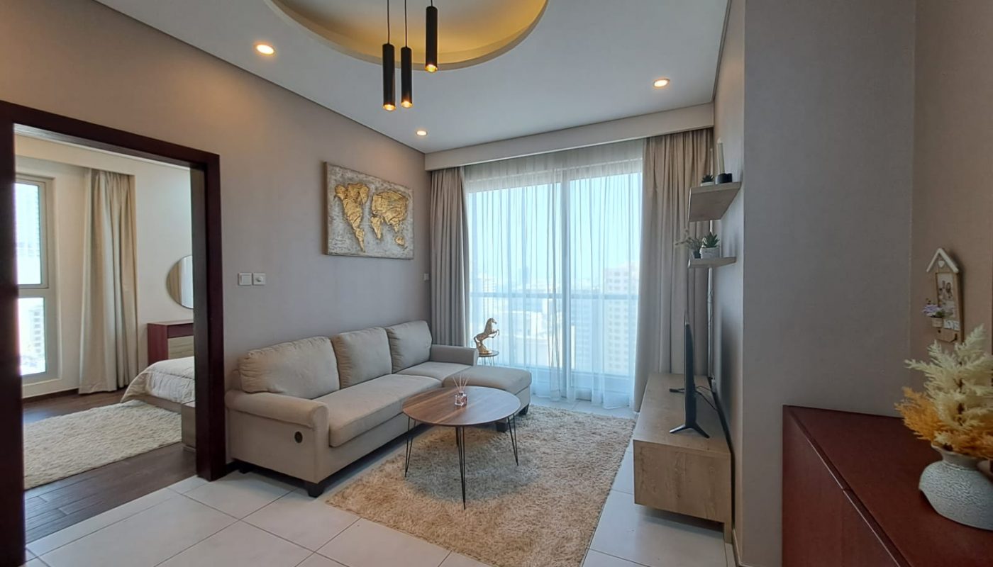 A modern living room with a beige sofa, round coffee table, wall-mounted TV, large window with curtains, and an adjoining bedroom visible through an open doorway.