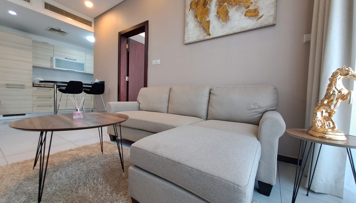 A modern living room with a beige sectional sofa, round wooden tables, a shaggy rug, and a gold horse sculpture; kitchen and barstools visible in the background.