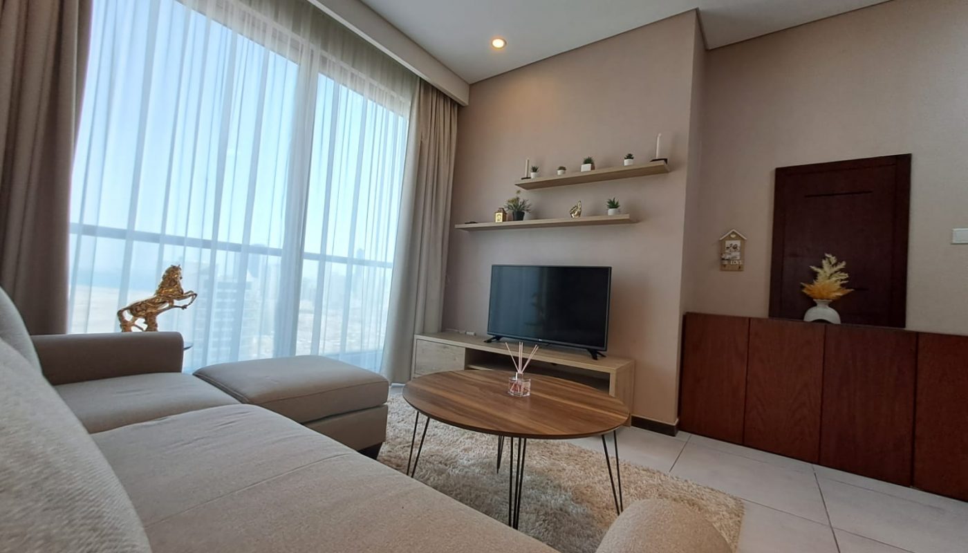 A modern living room with a beige sofa, round wooden coffee table, TV on a stand, wall shelves, large window with sheer curtains, and neutral decor.