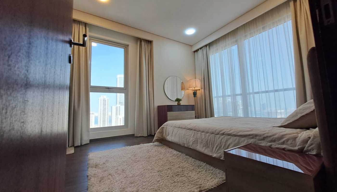 A modern bedroom with a beige bed, white rug, wooden furniture, large windows with sheer curtains, a round mirror, and a city view.
