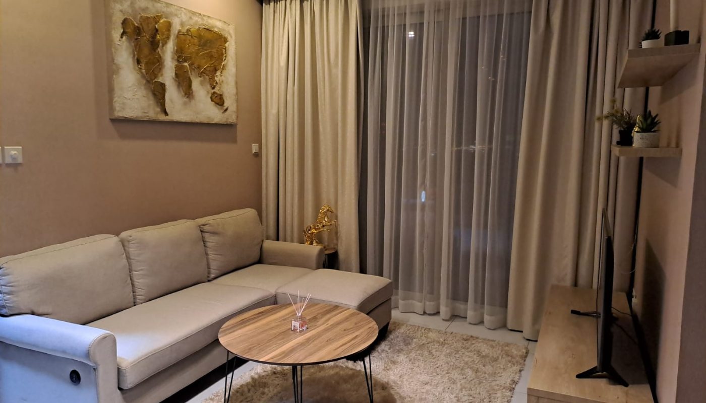 A modern living room with a beige sofa, round wooden coffee table, TV on a stand, wall art, and floor-to-ceiling curtains covering a large window.
