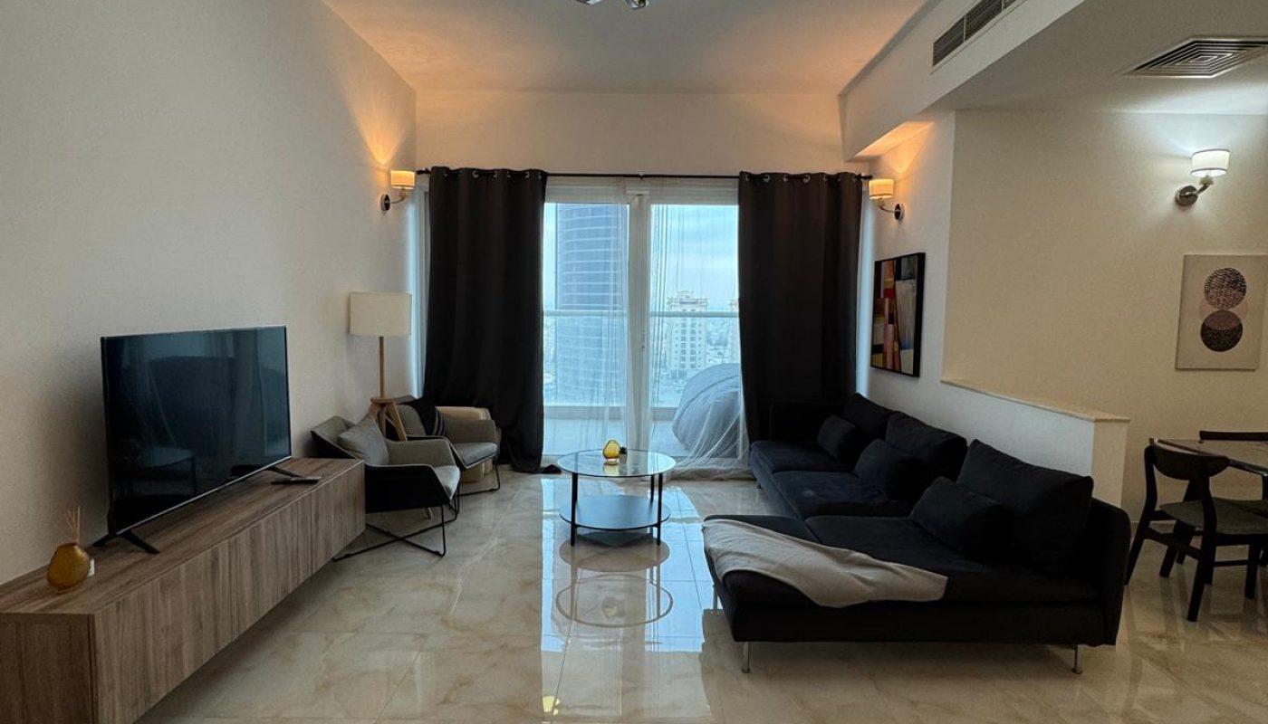 Modern living room with tiled floors, black sofa, TV on a wooden stand, armchair, glass coffee table, and large windows with dark curtains. Dining area visible to the right.
