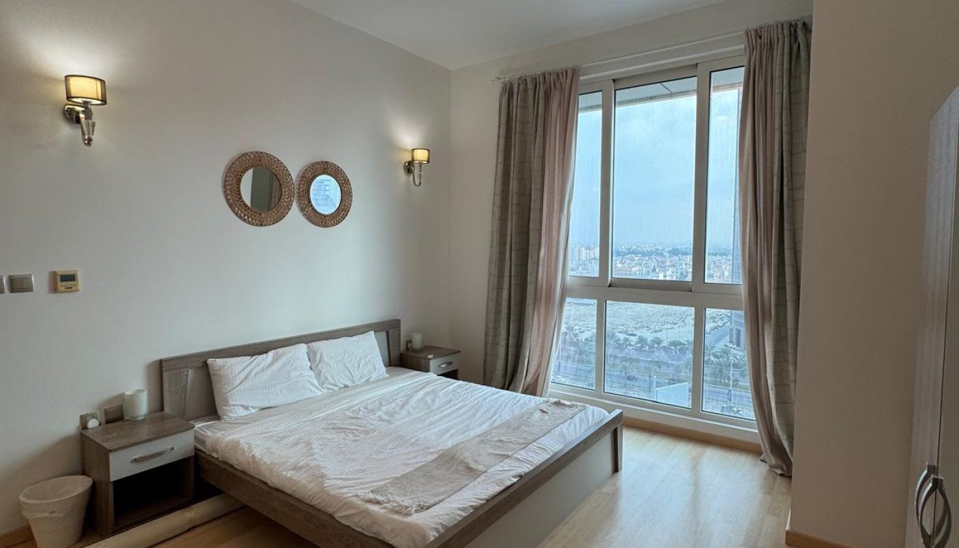 A minimalist bedroom with a double bed, bedside tables, wall mirrors, wooden flooring, large window with curtains, and a ceiling light fixture.