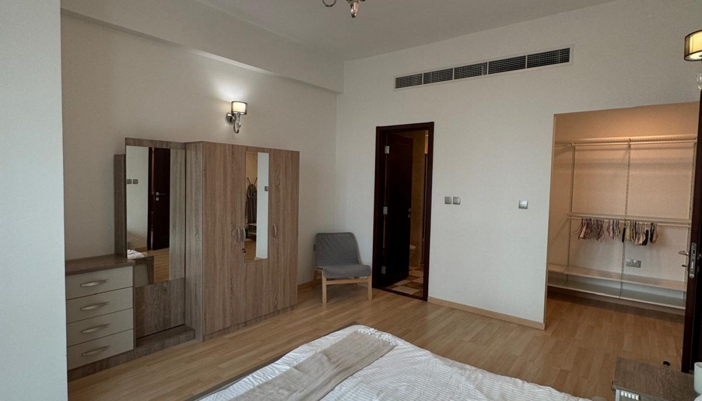 A bedroom with a double bed, wooden wardrobe, chair, dresser, open walk-in closet, and overhead chandelier, featuring light-colored flooring and walls.