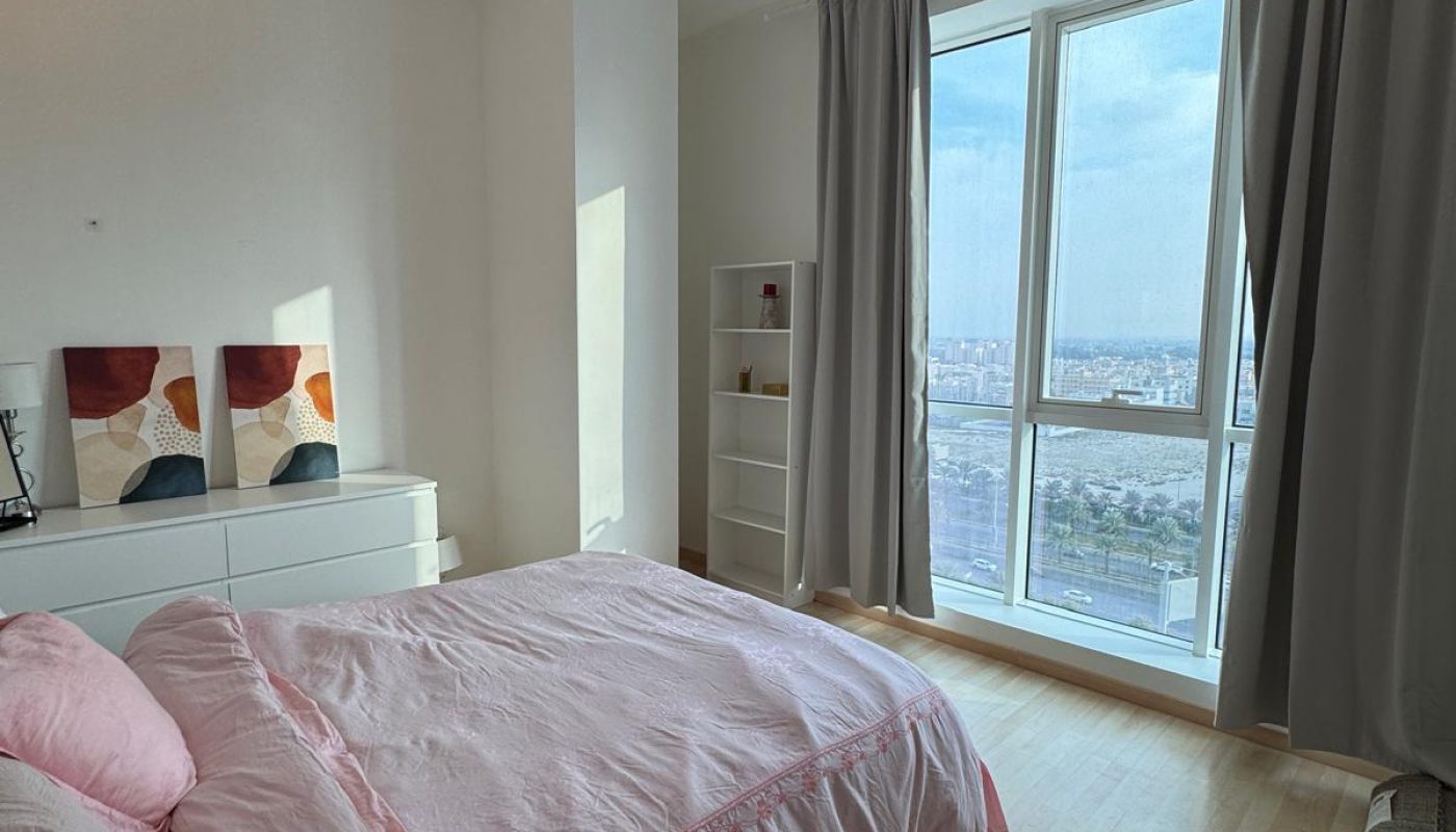 A tidy bedroom with a pink bedspread, white dresser, two abstract paintings, a small bookshelf, large window with gray curtains, and a city view.